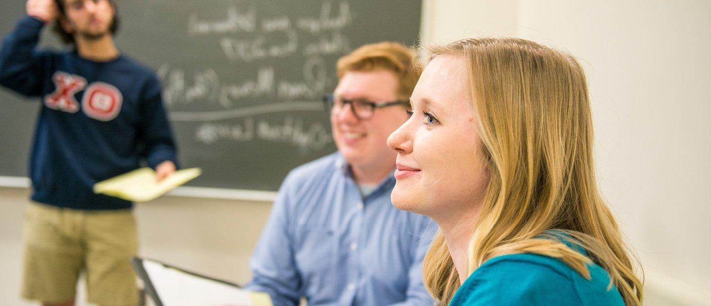 A young man and woman seated, looking towards the front a classroom while a young man stands behind them by a chalkboard.